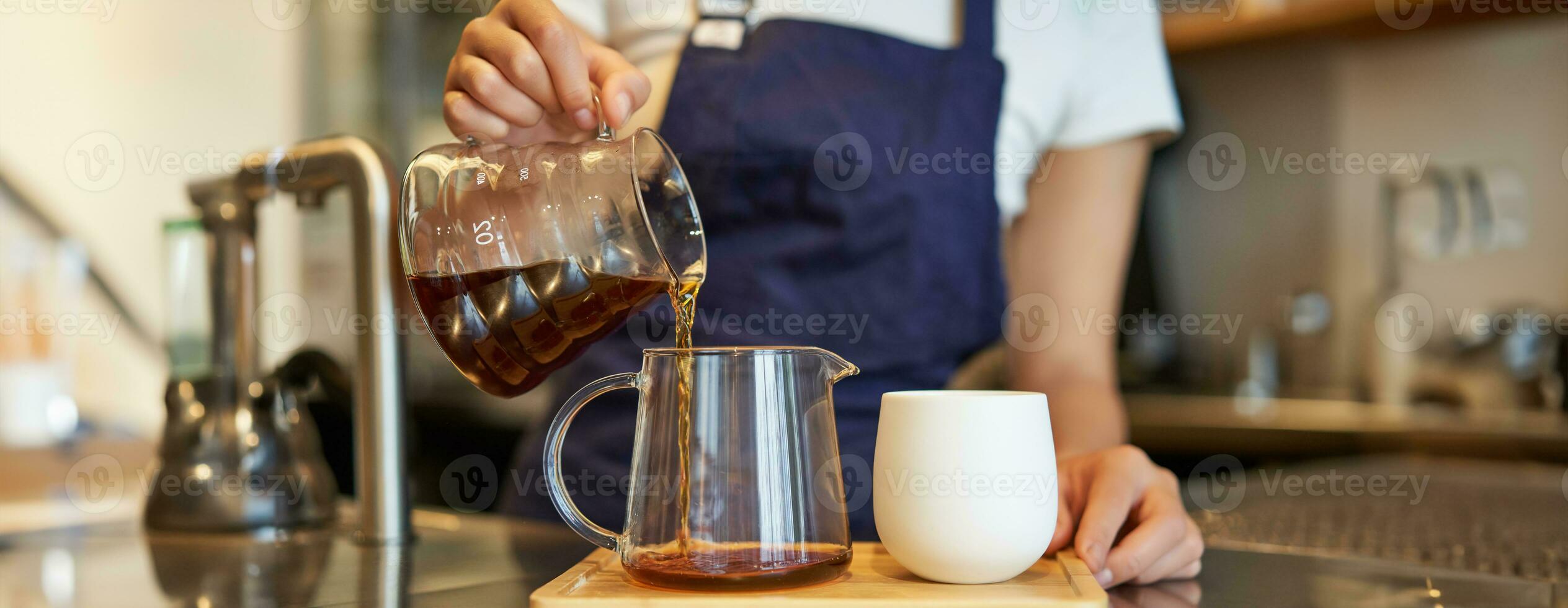Close up of glass jar and a cup standing on cafe counter, barista pouring filter coffee and preparing order photo