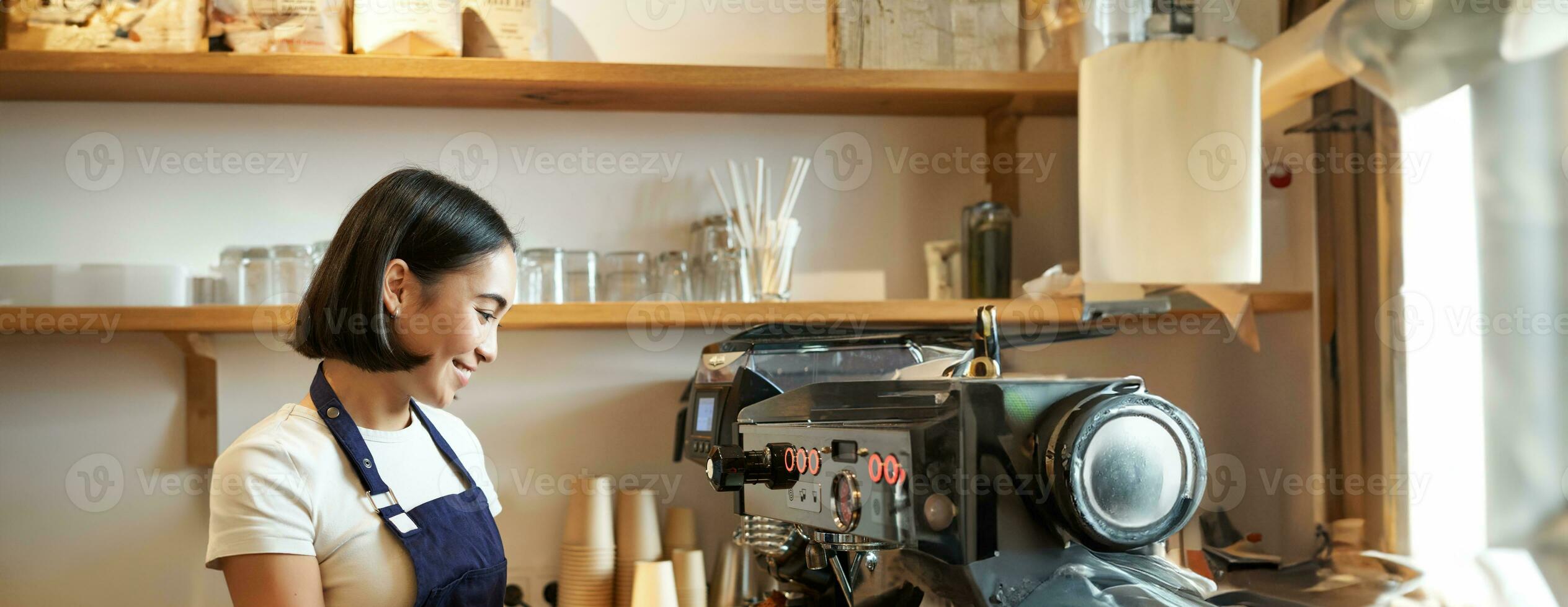 contento hermosa asiático chica, barista en delantal trabajos con café máquina detrás encimera, en pie en café y haciendo capuchino foto