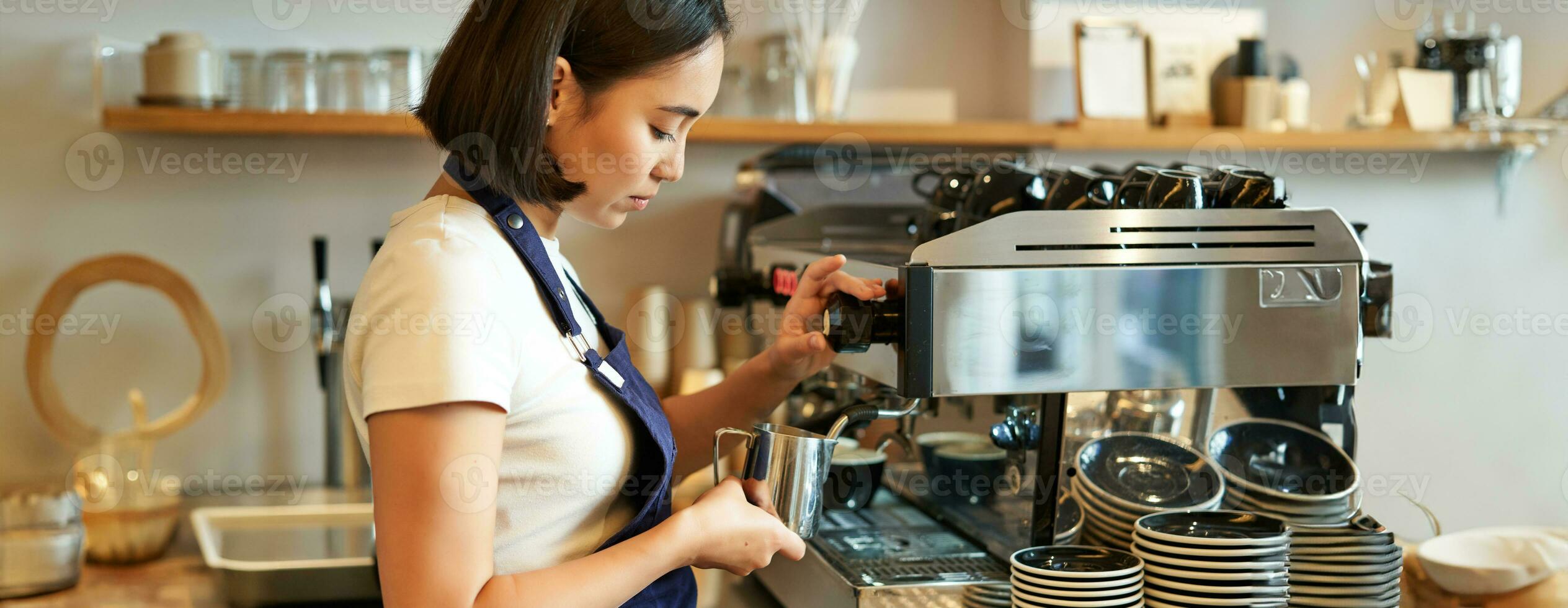 Smiling young asian barista girl, using coffee machine steamer