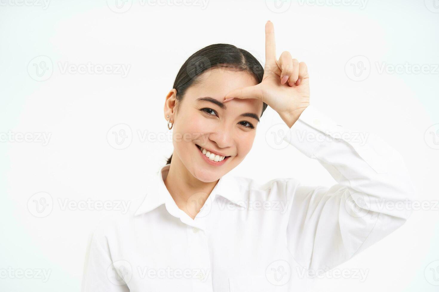 Asian woman shows L letter, loser on forehead and smiling, winner mocks person who failed, stands over white background photo