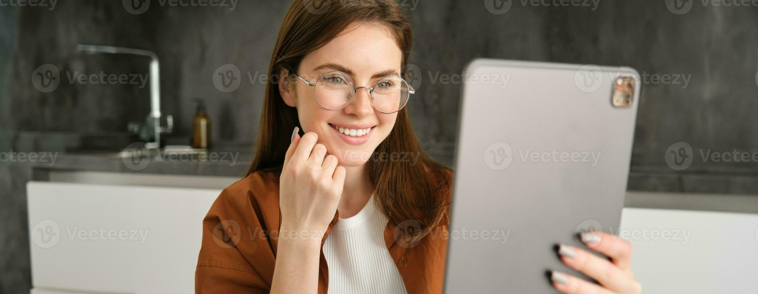 retrato de hermosa joven mujer en anteojos, mirando a digital tableta. mujer de negocios trabajos en remoto, persona de libre dedicación leyendo en artilugio, sentado en cocina foto