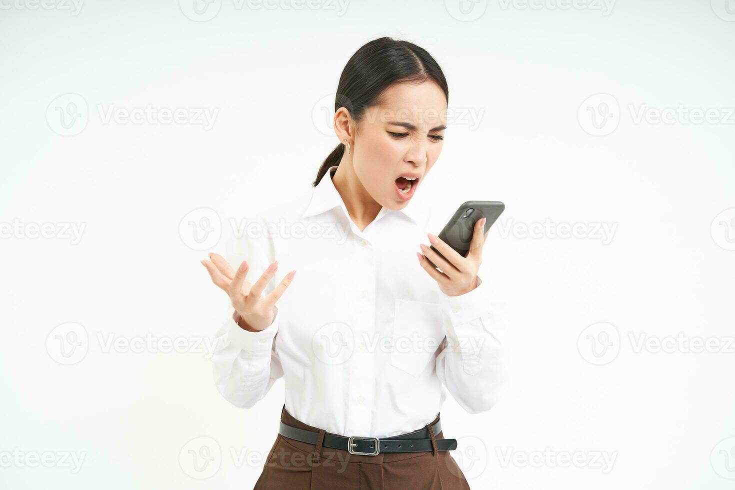Angry asian businesswoman shouts at her mobile phone with frustrated face expression, stands isolated on white background photo