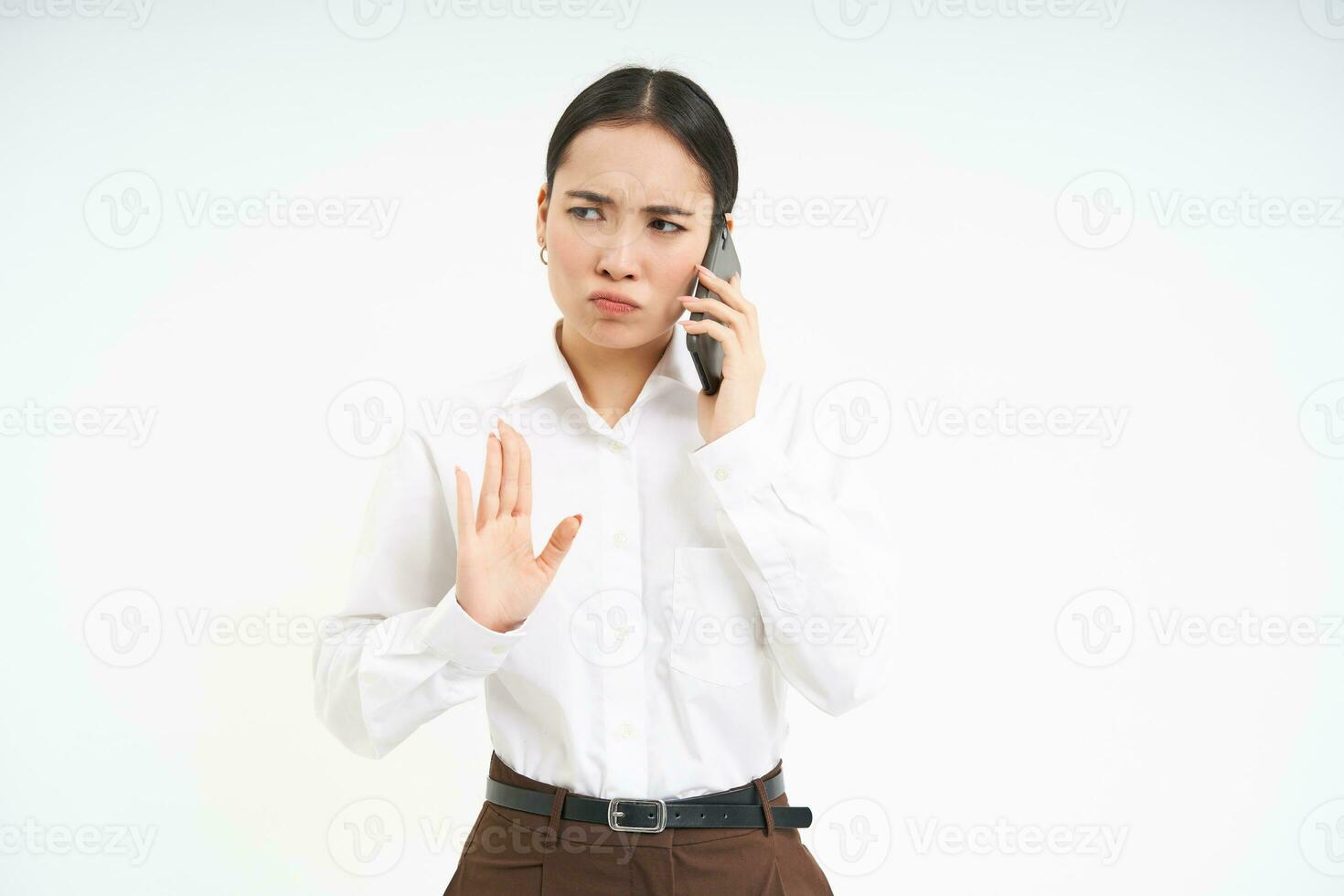 Portrait of serious korean woman, business professional shows stop sign, talks on mobile phone with disapproval, frowns upset, stands over white background photo