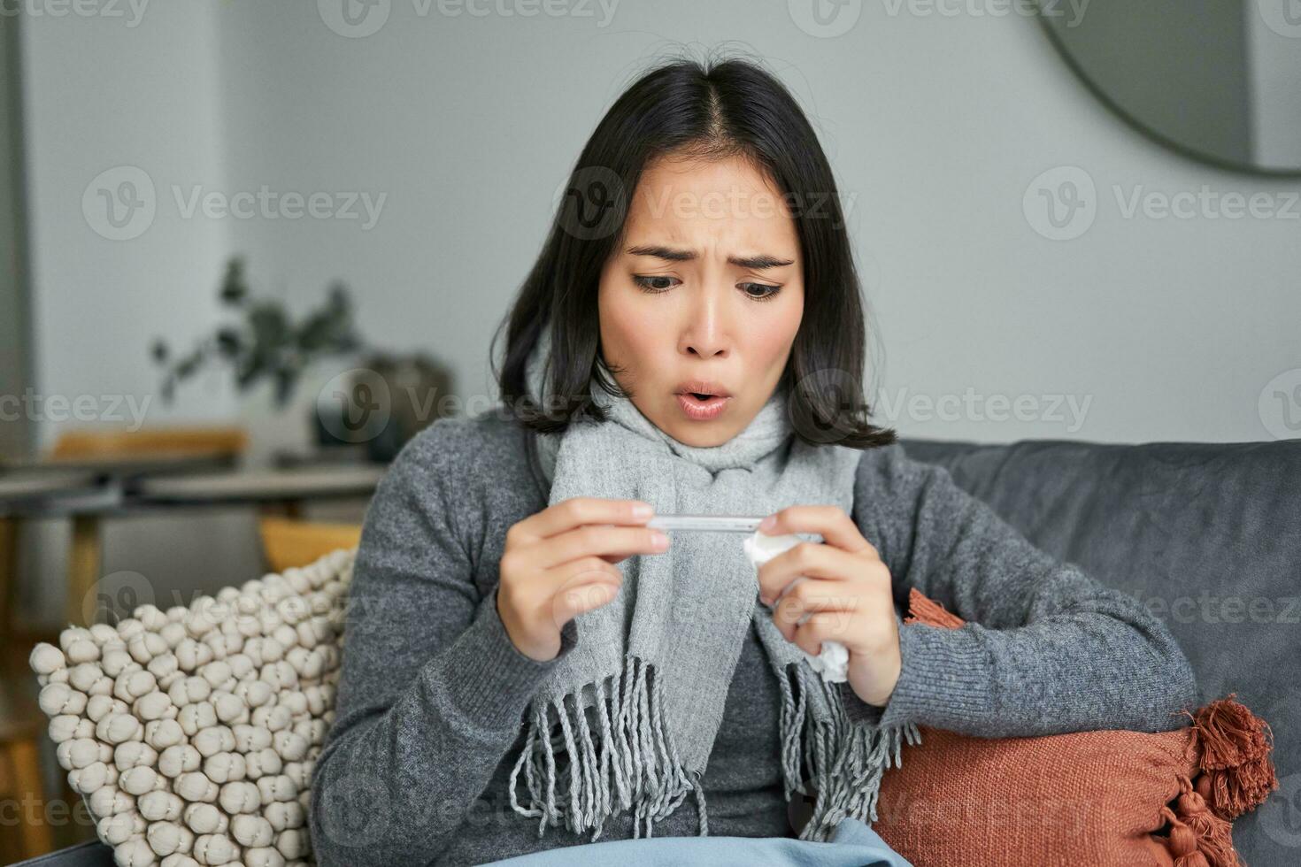 Shocked woman looks concerned at her thermometer, catching cold, has fever, sitting on sick leave at home photo