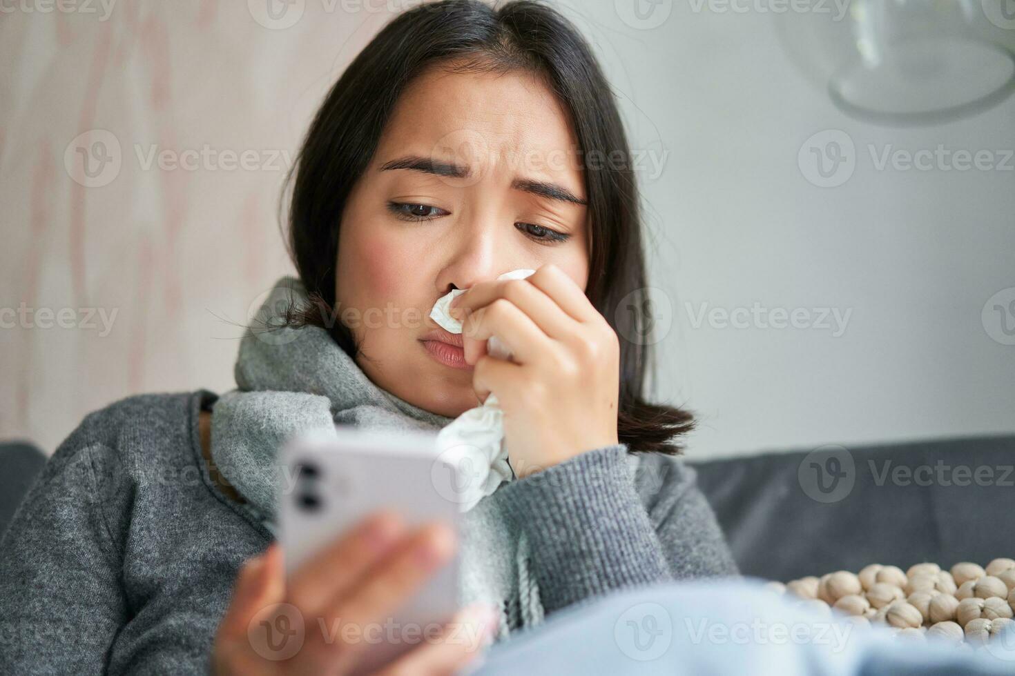 Ill korean woman sitting at home, sneezing, caught cold, reading news on mobile phone, freezing at her home because of heating problems photo