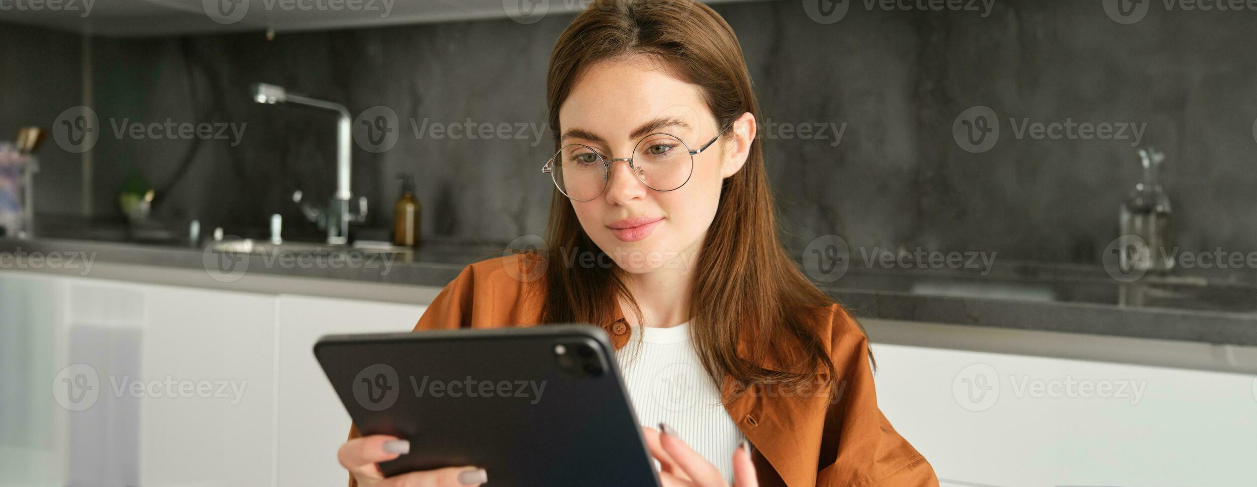 Portrait of busy young woman, self-employed entrepreneur sitting at home with digital tablet, reading documents. Student working on project remotely, holding gadget, studying photo