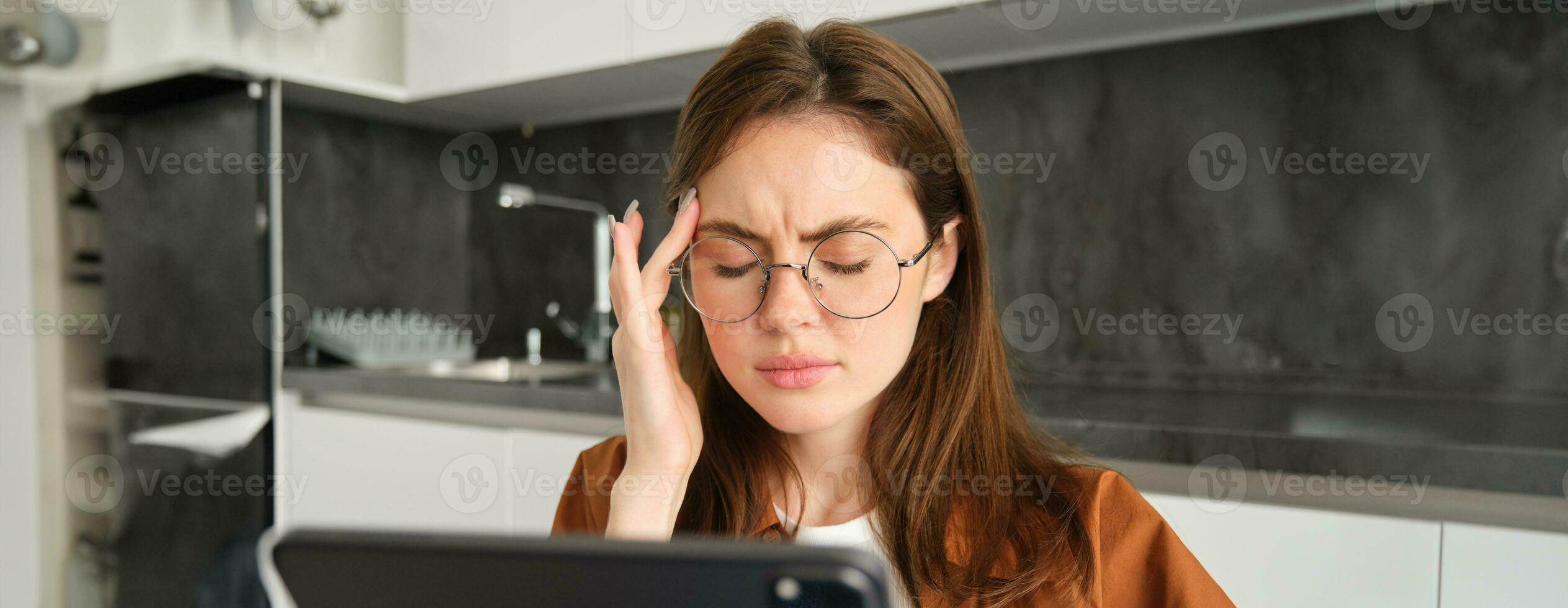 Close up portrait of brunette woman in glasses, grimacing from painful migraine or headache, feels unwell while reads digital tablet, works from home in kitchen photo