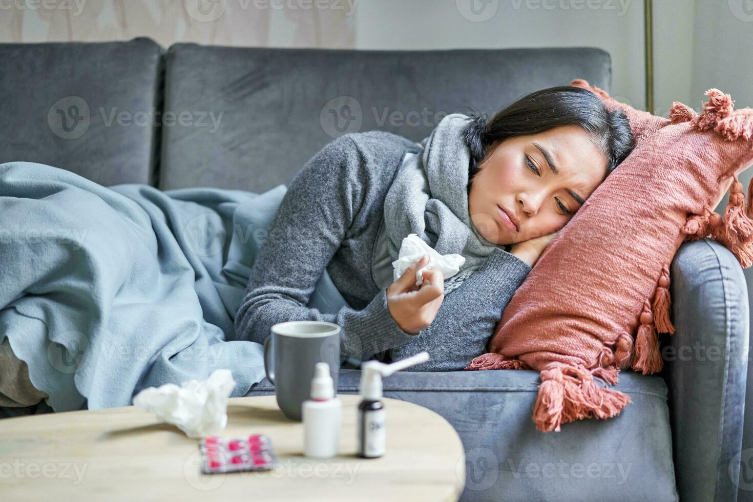 Sick sad korean woman lying on sofa, feeling unwell, catching cold, flu and temperature, looking upset, taking medication photo