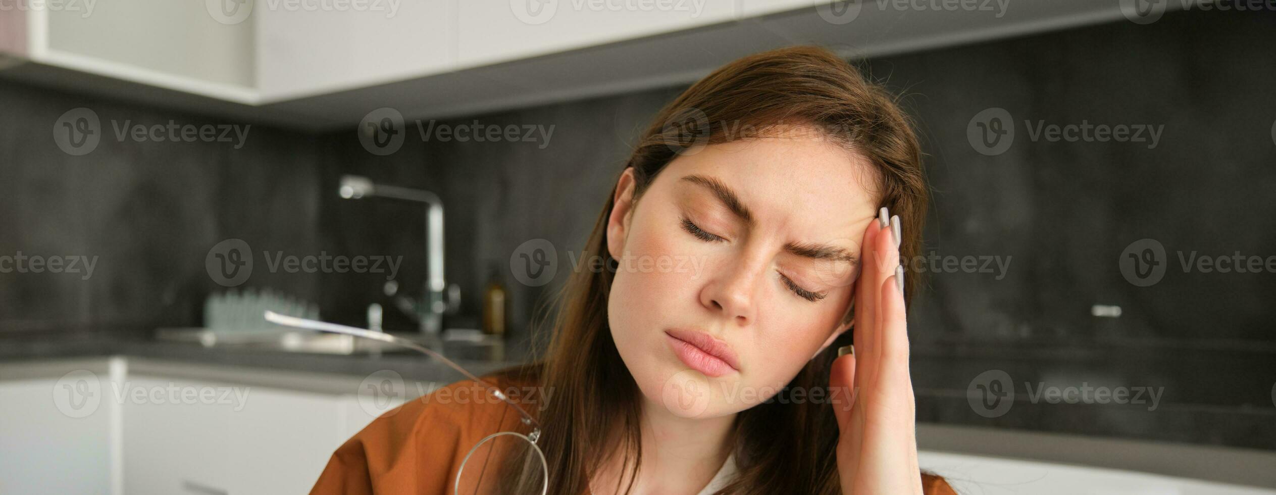 Close up portrait of tired young woman, wife sitting in kitchen and touching head, has headache, massaging temple and frowning from painful migraine photo