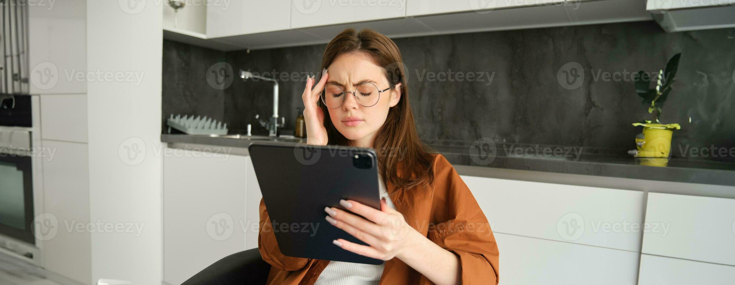 Portrait of woman freelancer, wearing glasses, holding digital tablet, looking tired, has migraine or headache, reading e-book. Freelance worker sits at home with gadget photo