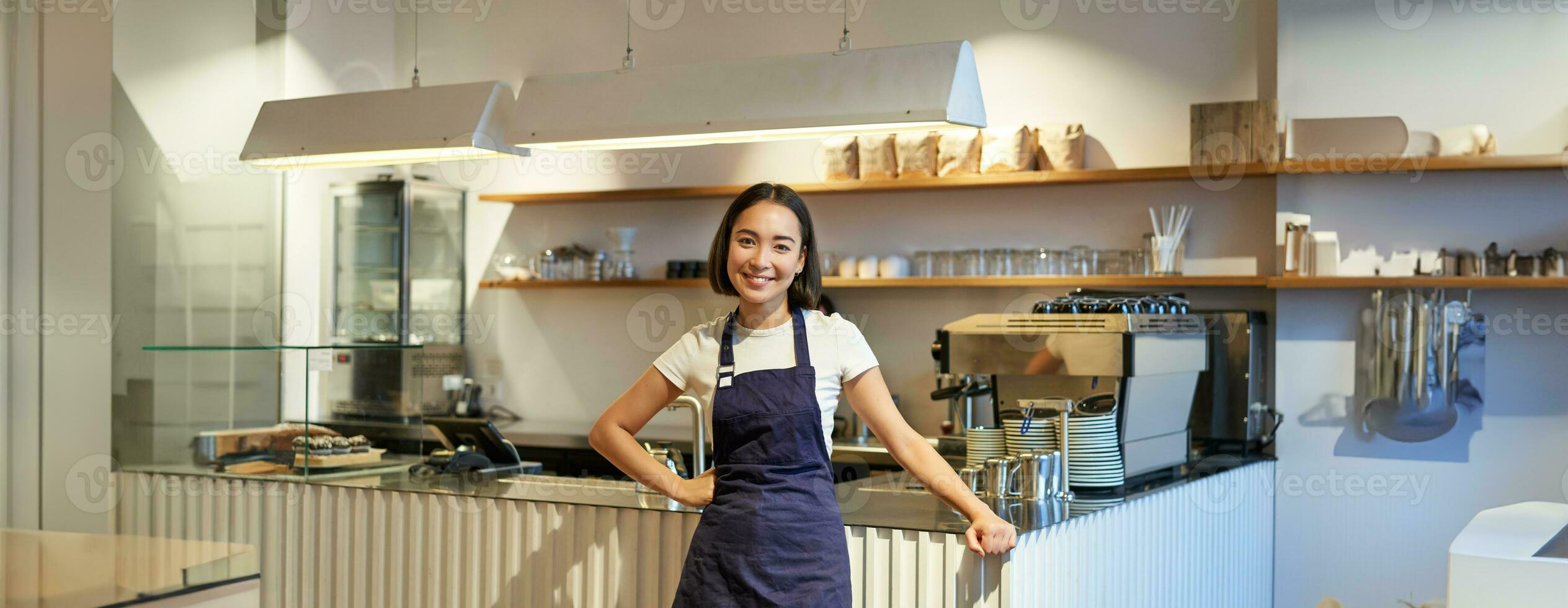 retrato de linda asiático mujer barista, café personal en pie cerca mostrador con café máquina, vistiendo delantal, sonriente a cámara foto