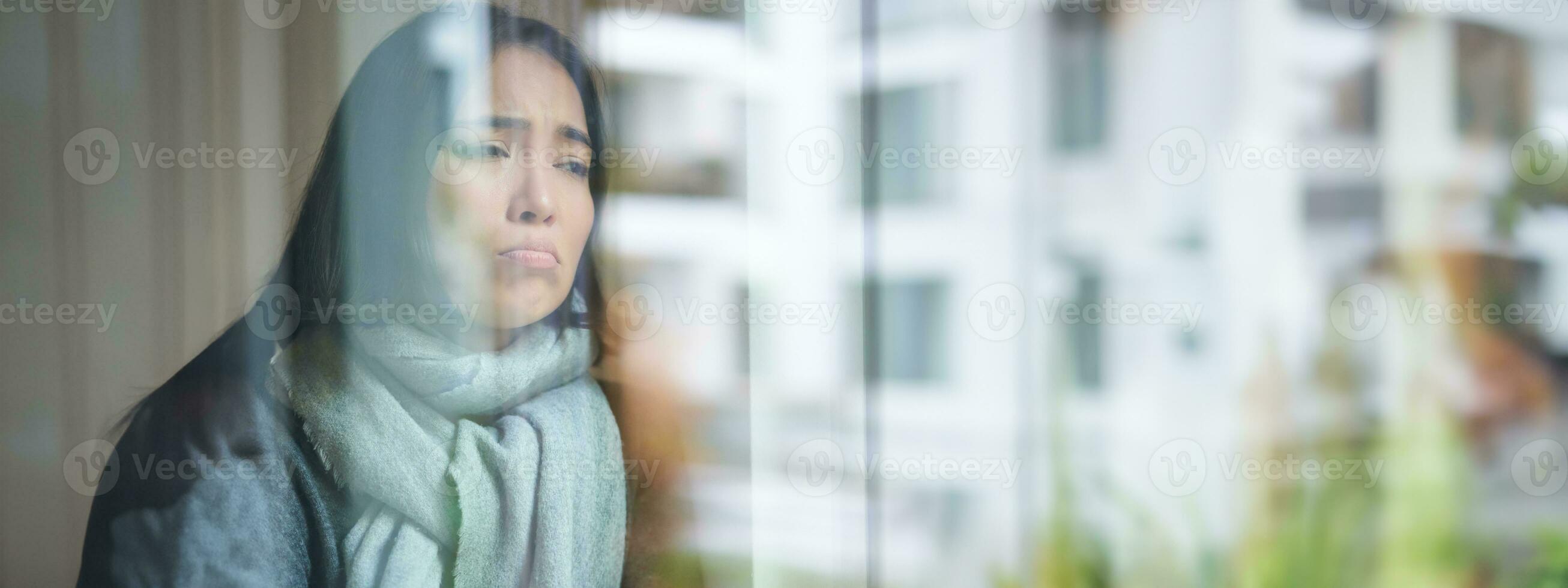 Portrait of sad ill korean woman looking out of the window, staying at home due to coronavirus, being sick and cant leave house photo