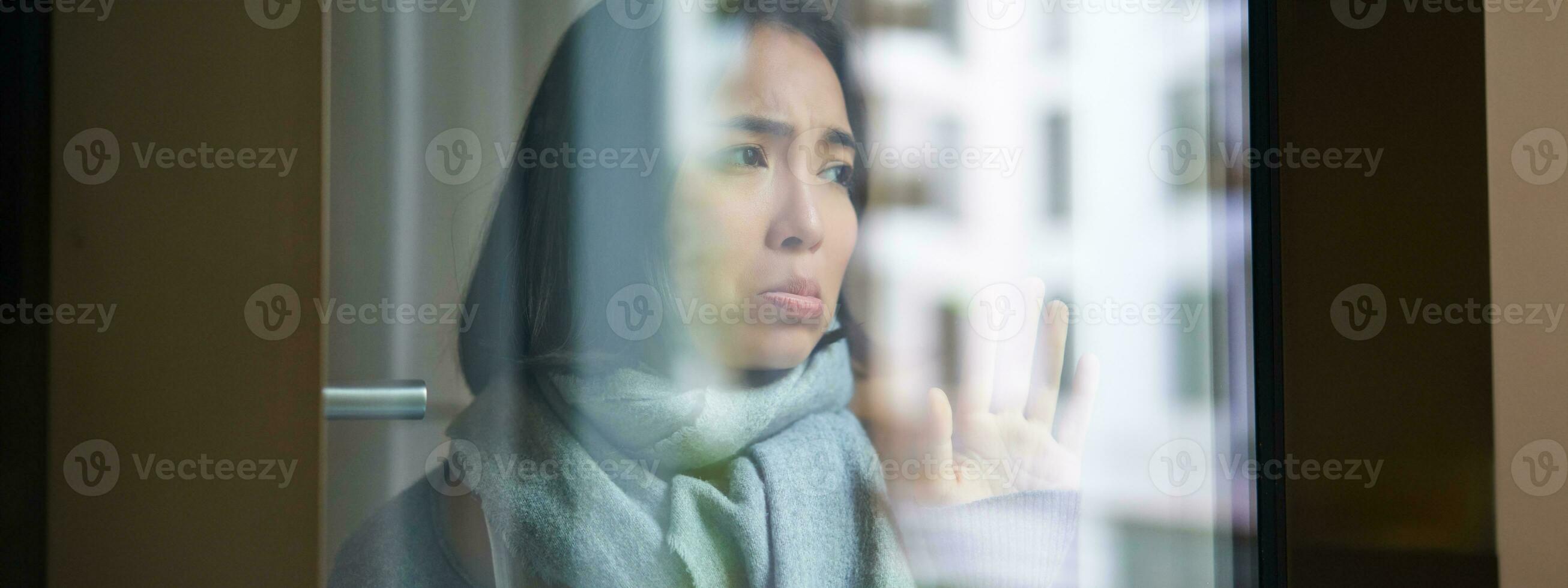 Portrait of sad ill korean woman looking out of the window, staying at home due to coronavirus, being sick and cant leave house photo