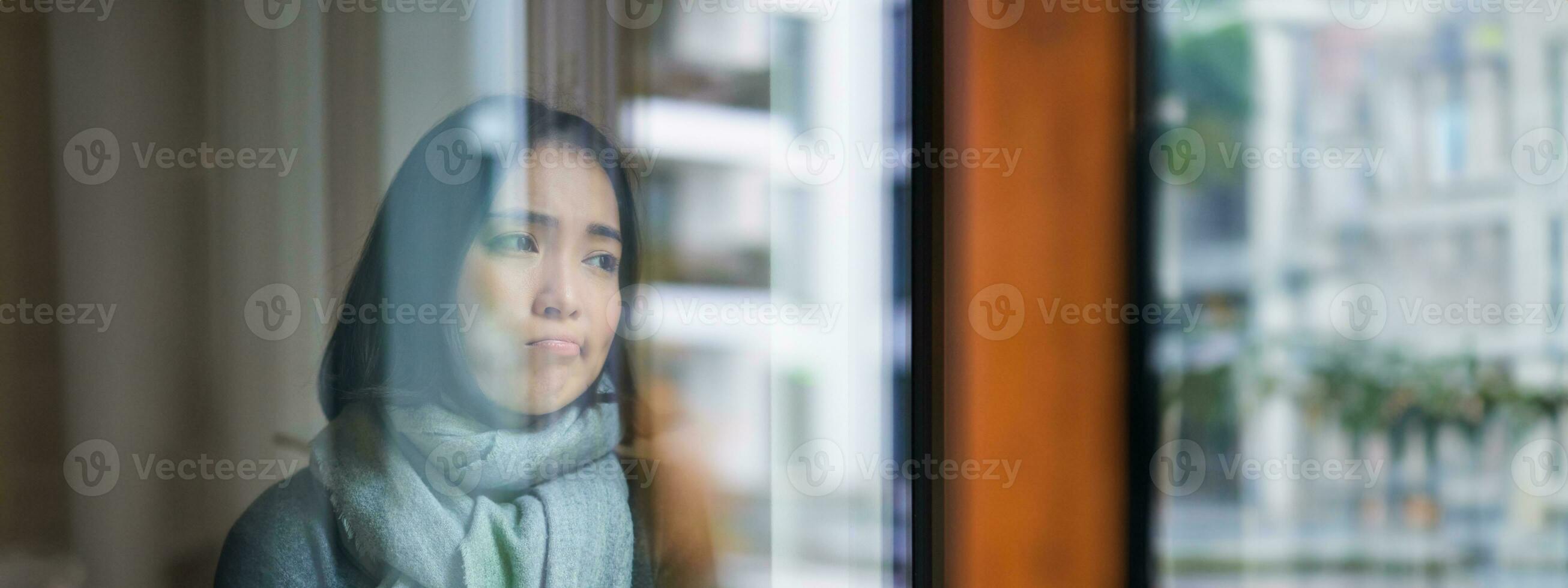 Portrait of sad ill korean woman looking out of the window, staying at home due to coronavirus, being sick and cant leave house photo