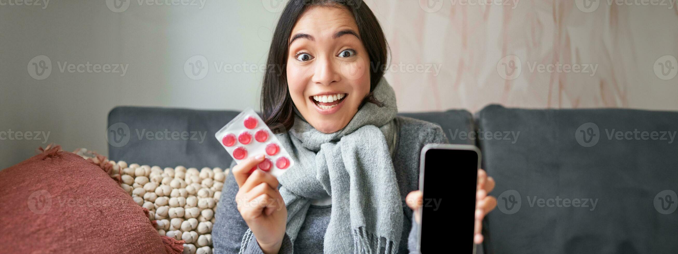 Smiling korean girl shows smartphone screen, medication in hands, feeling sick and staying at home, using online GP doctor app while on sick leave photo