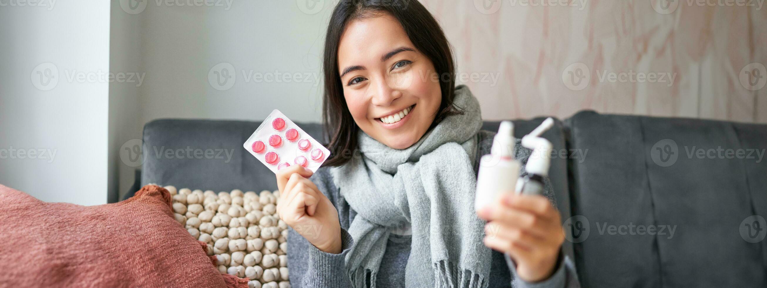 Portrait of happy smiling asian girl showing medication, sore throat spray and drugs from flu or cold, staying at home, wearing warm clothes photo