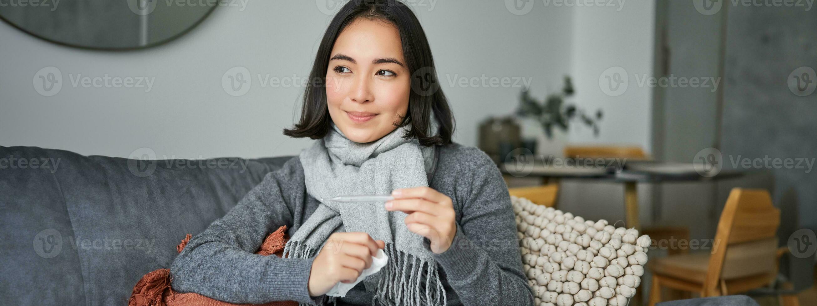 Smiling asian woman holding thermometer and looking pleased, feeling better after cold, got rid of fever, has normal temperature photo
