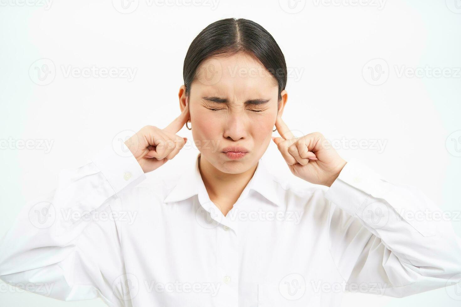 Annoyed young asian corporate worker, lady screams and shuts her ears with fingers, stands frustrated against white studio background photo