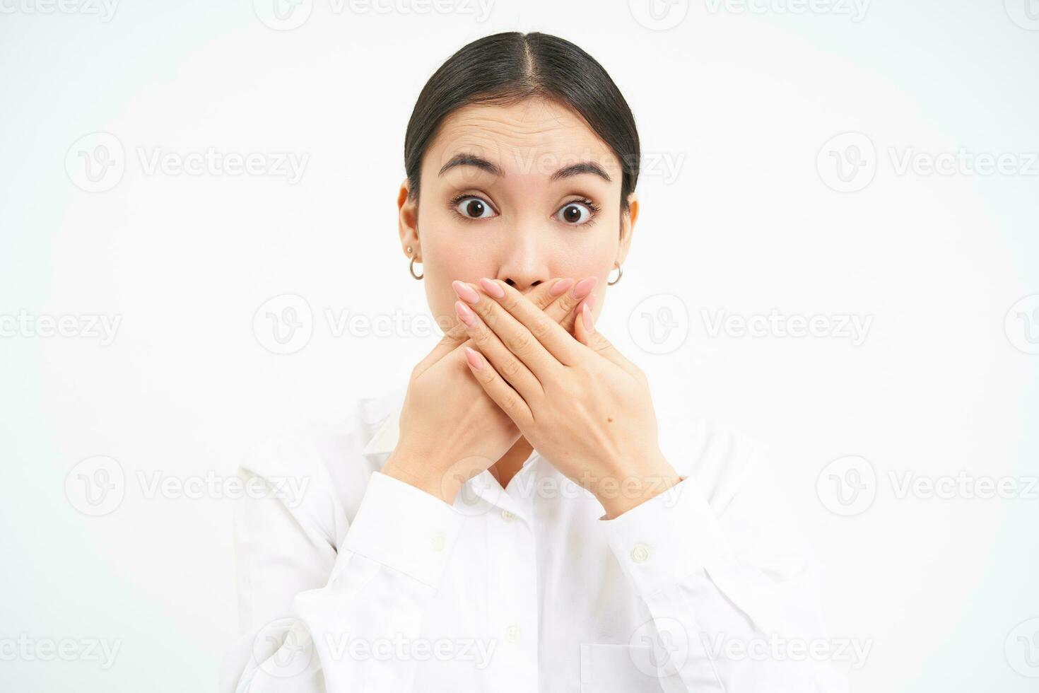 Portrait of korean business woman, shuts her mouth, holds hands on lips with shocked, surprised face expression, stands over white background photo