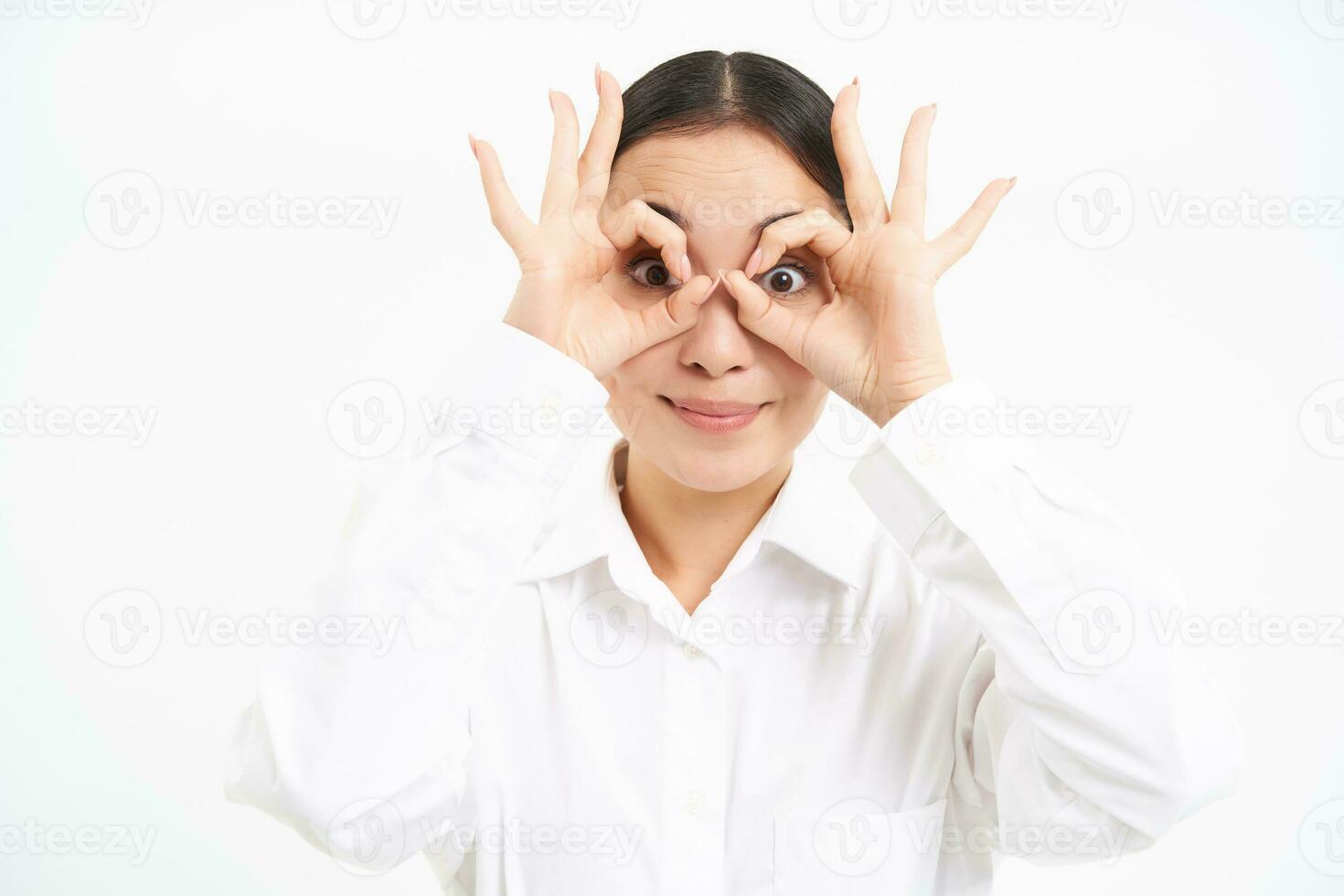 Business people. Happy asian girl looks through finger glasses, stares with surprised face, stands isolated on white background photo