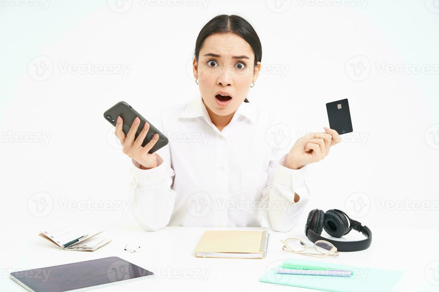 Portrait of asian woman sits at table, holds credit card and smartphone, looks upset, disappointed by prices, white background photo