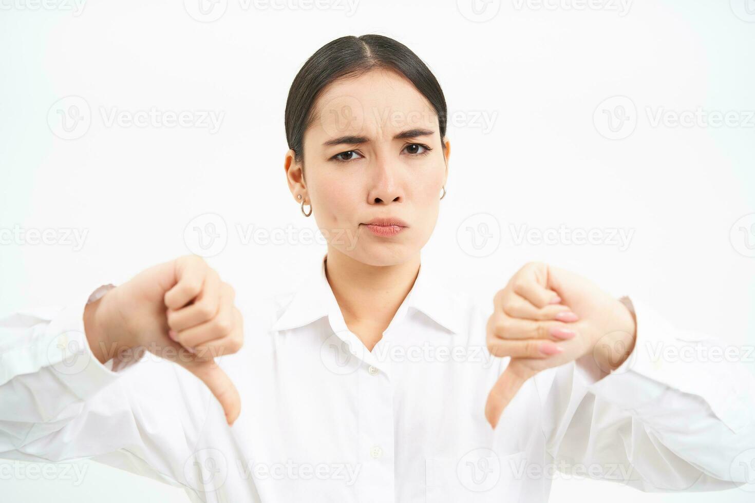 Portrait of disappointed japanese woman, shows thumbs down, frowns and shakes head with disapproval, stands over white background photo