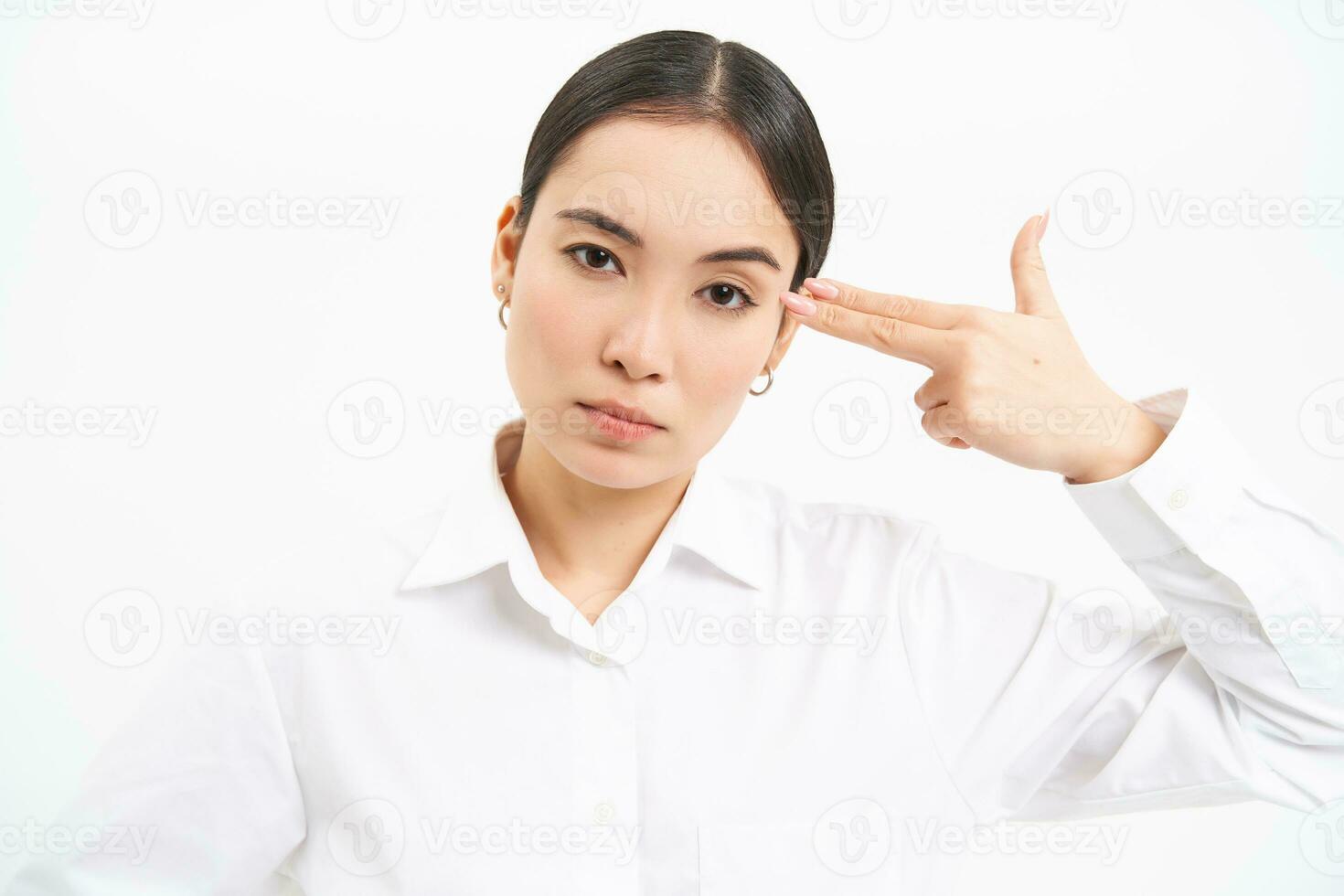 irritado y cansado asiático mujer de negocios, muestra pistola gesto en cabeza, mira molesto y tenso, soportes terminado blanco antecedentes foto