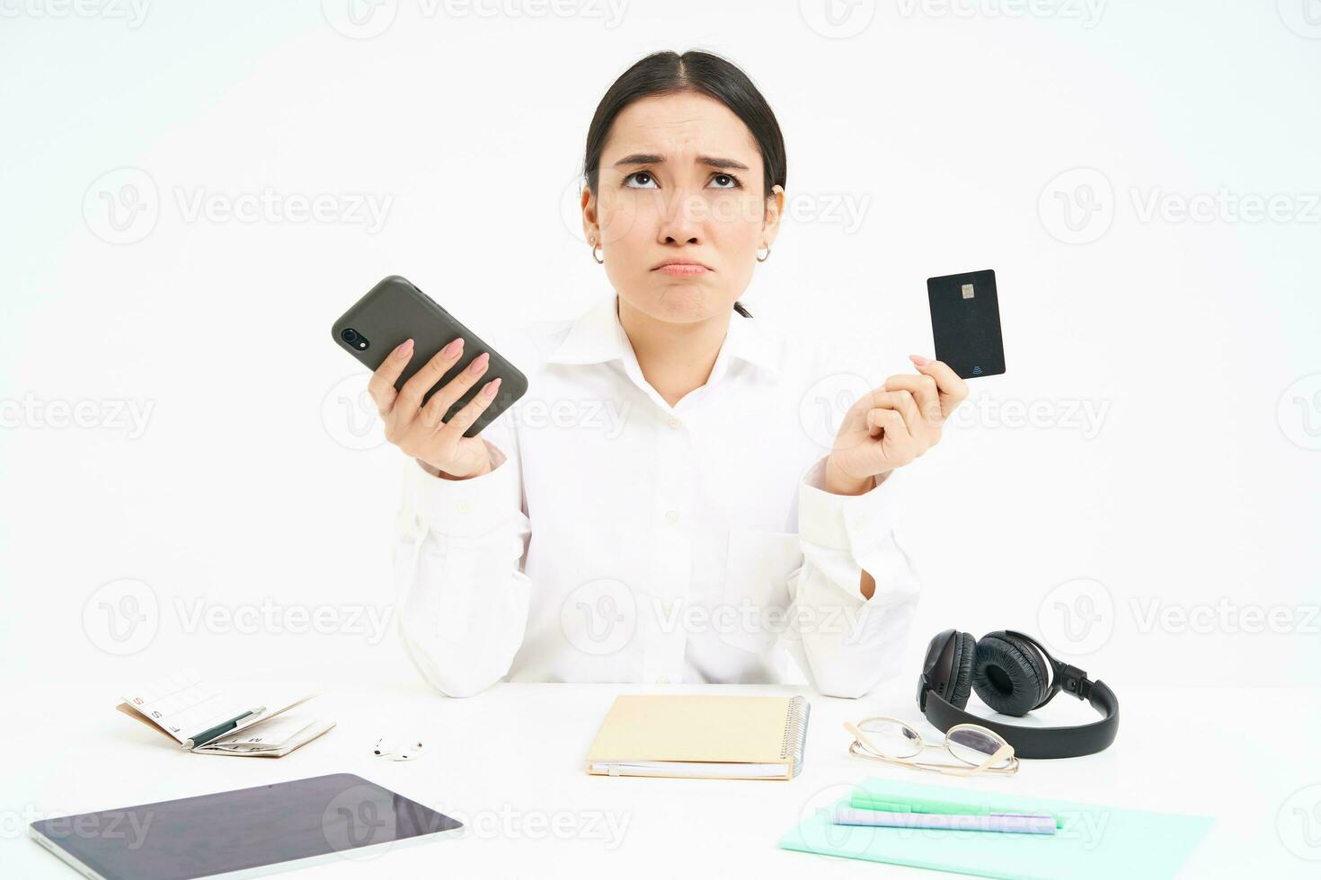 Portrait of asian woman sits at table, holds credit card and smartphone, looks upset, disappointed by prices, white background photo