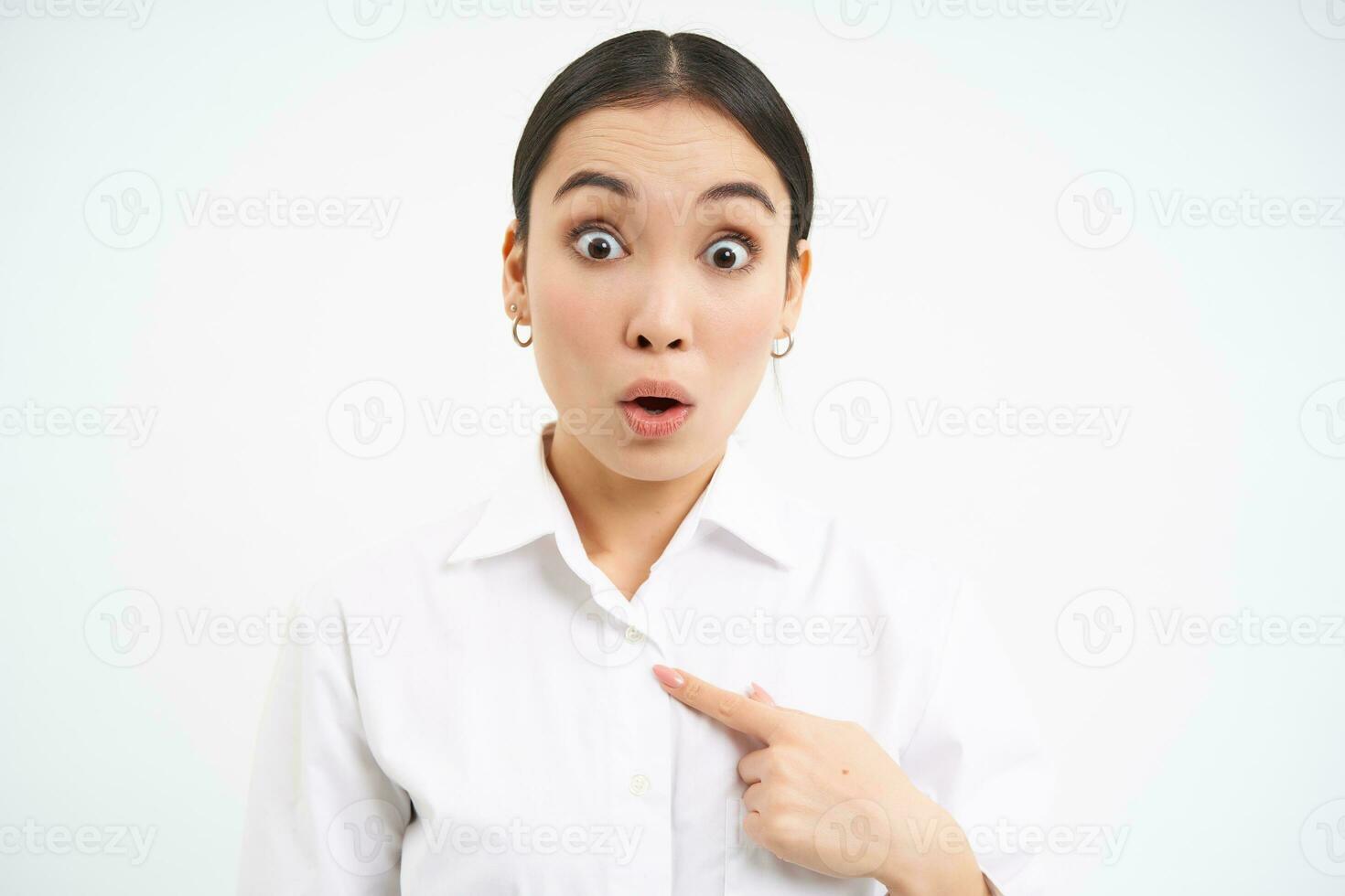 Portrait of asian businesswoman looks surprised, points at herself with shocked face expression, stands over white background photo