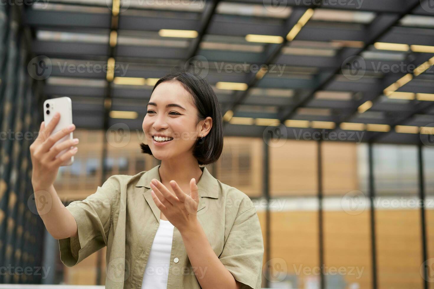 Happy asian girl shows something behind her during video call, demonstrating smth, standing on street photo