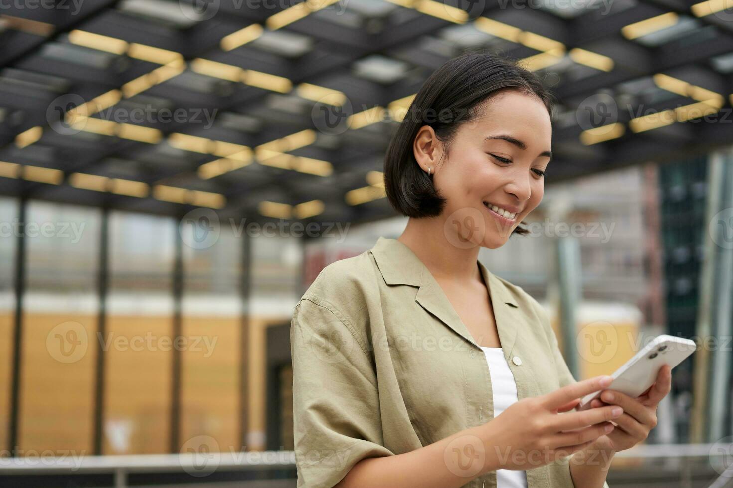 Technology people. Young smiling asian girl holding smartphone, using mobile app while standing on street, navigating around city photo