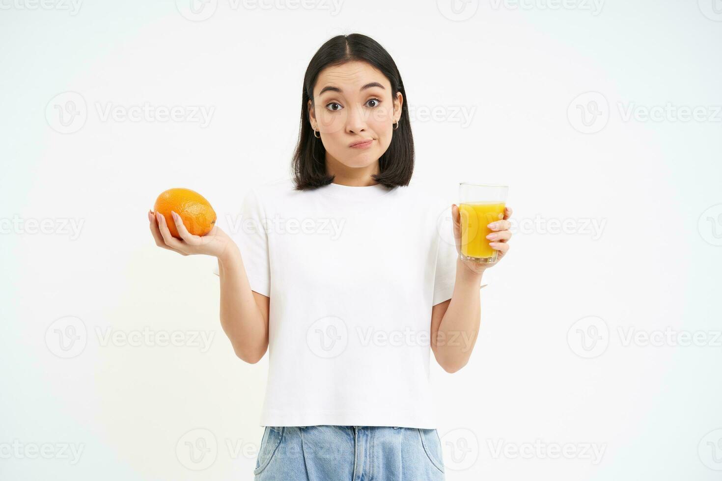 Indecisive young asian woman, holding orange and juice, shrugging, making choice, isolated on white background photo