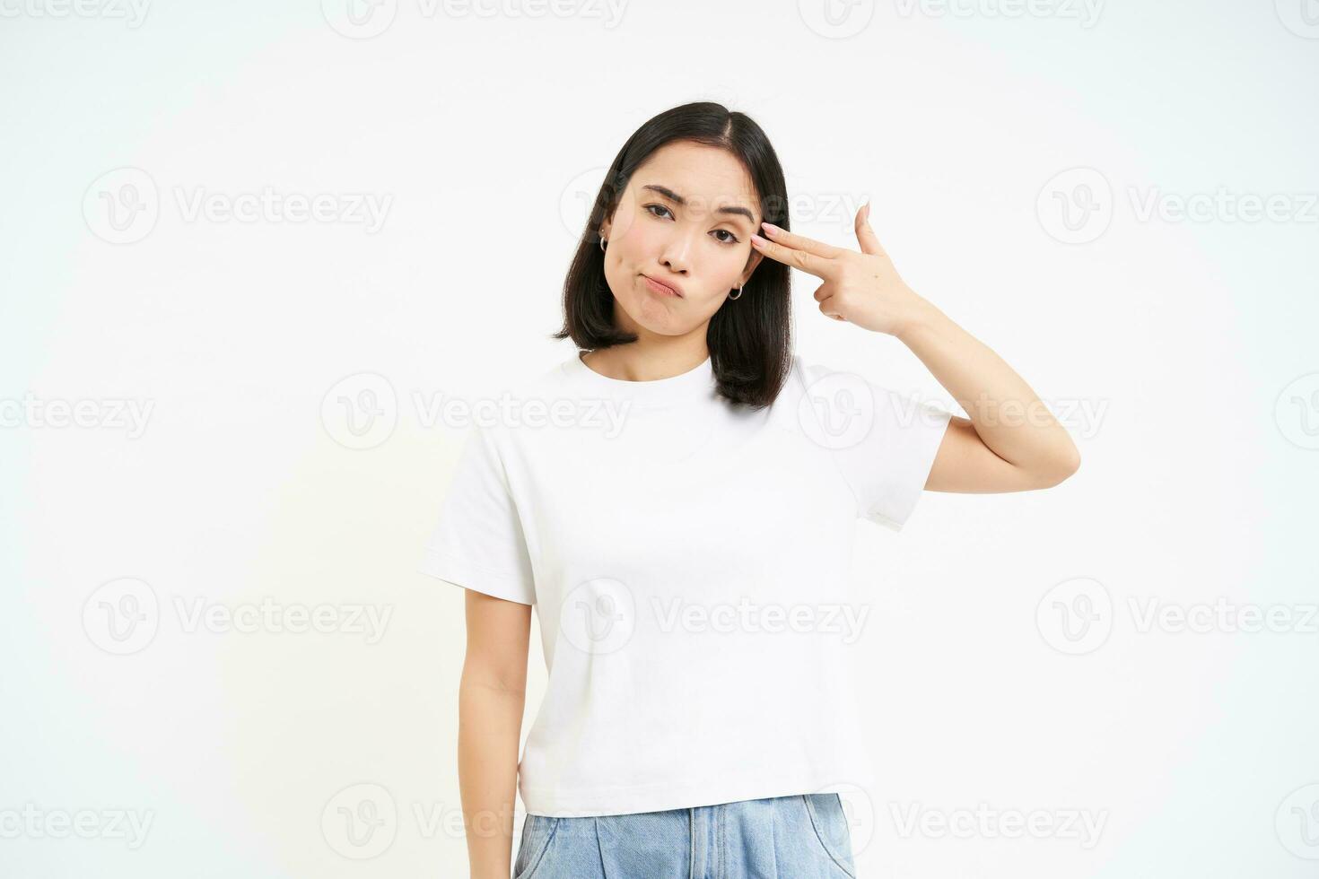 Annoyed asian woman, student shows finger pistol near head, looks bothered and tired, stands isolated on white background photo