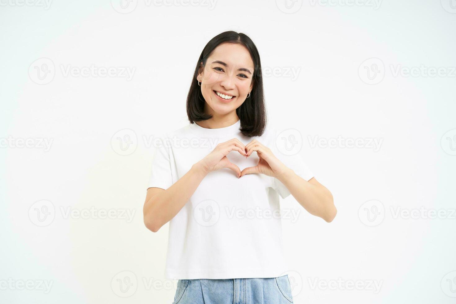Cute young asian woman, shows heart, love sign, looks with care and tenderness, white background photo