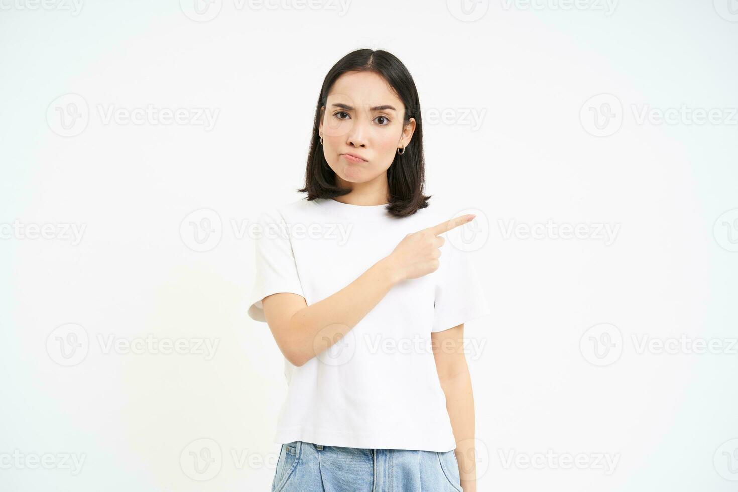Portrait of angry asian woman, pointing finger right with judgemental face, complaining and showing smth upsetting, white background photo