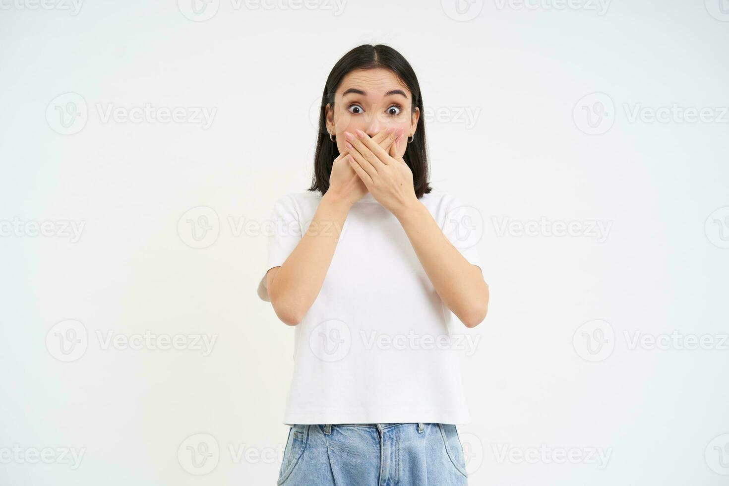 Portrait of girl with shocked face, holds hands on her mouth, stares speechless, startled by gossip, white background photo