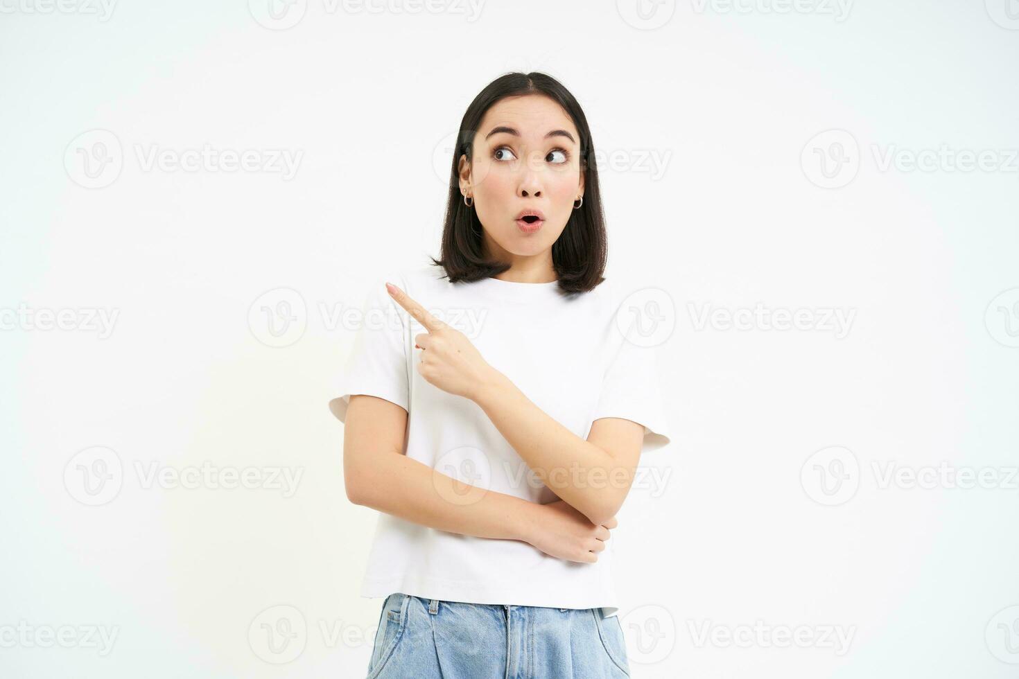Surprised korean girl, points left and looks with amazed face expression, stands in tshirt isolated on white background photo