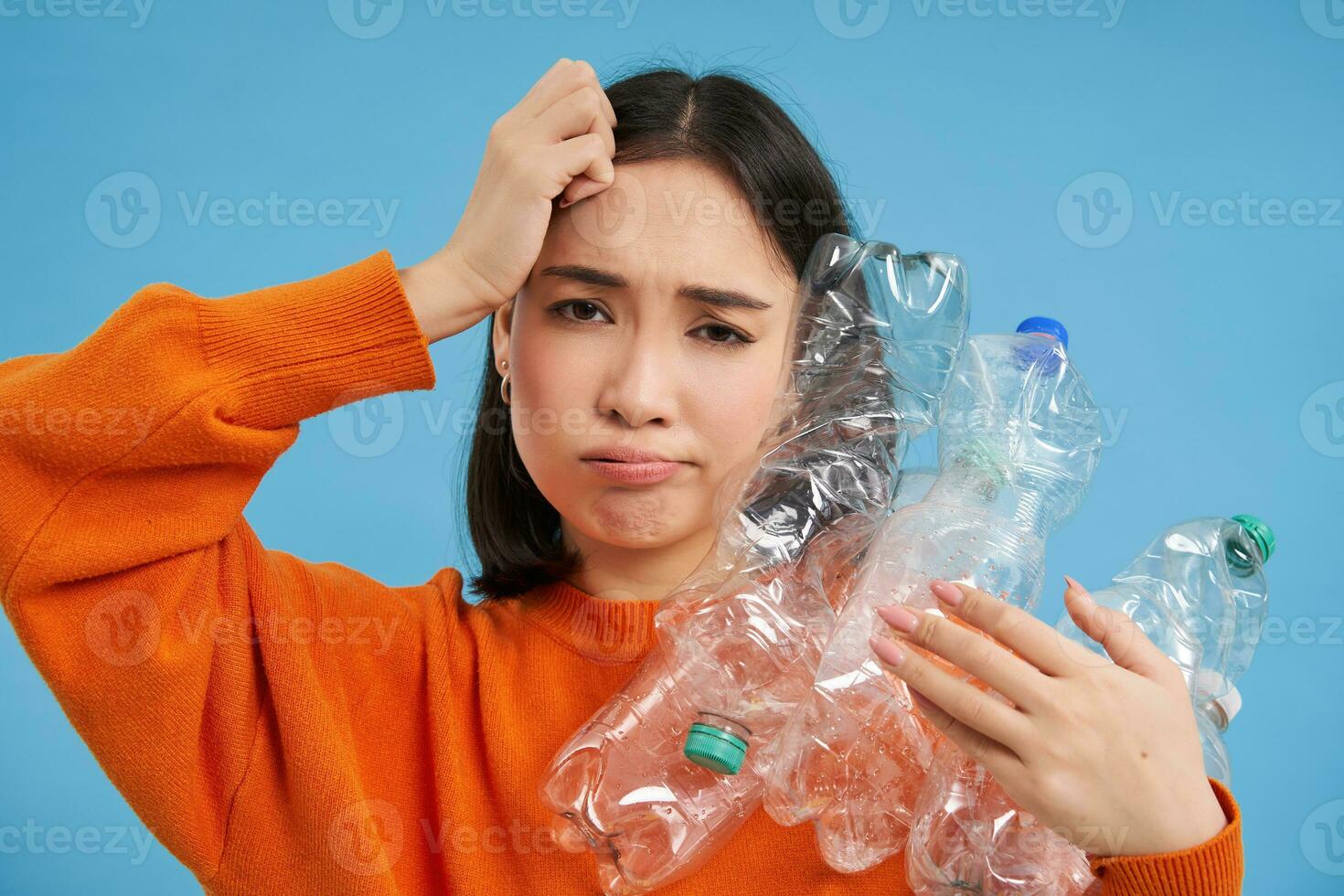 Troubled asian woman holding plastic bottles, trying find recycling center, blue background photo