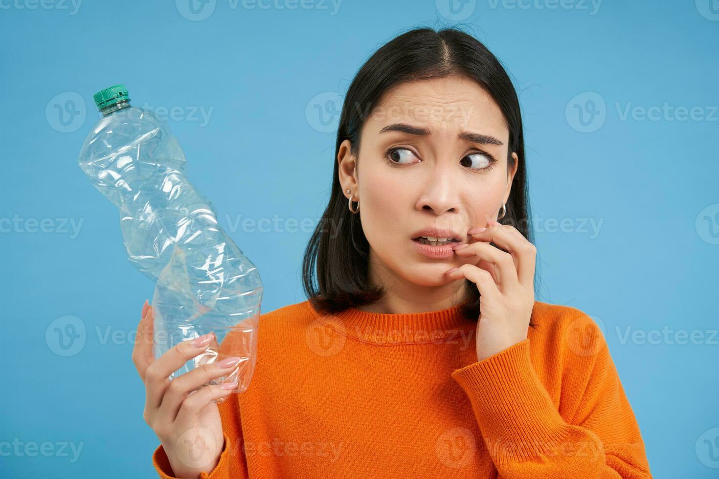 Portrait of girl looks scared at plastic bottle, worried about recycling, standing over blue background photo
