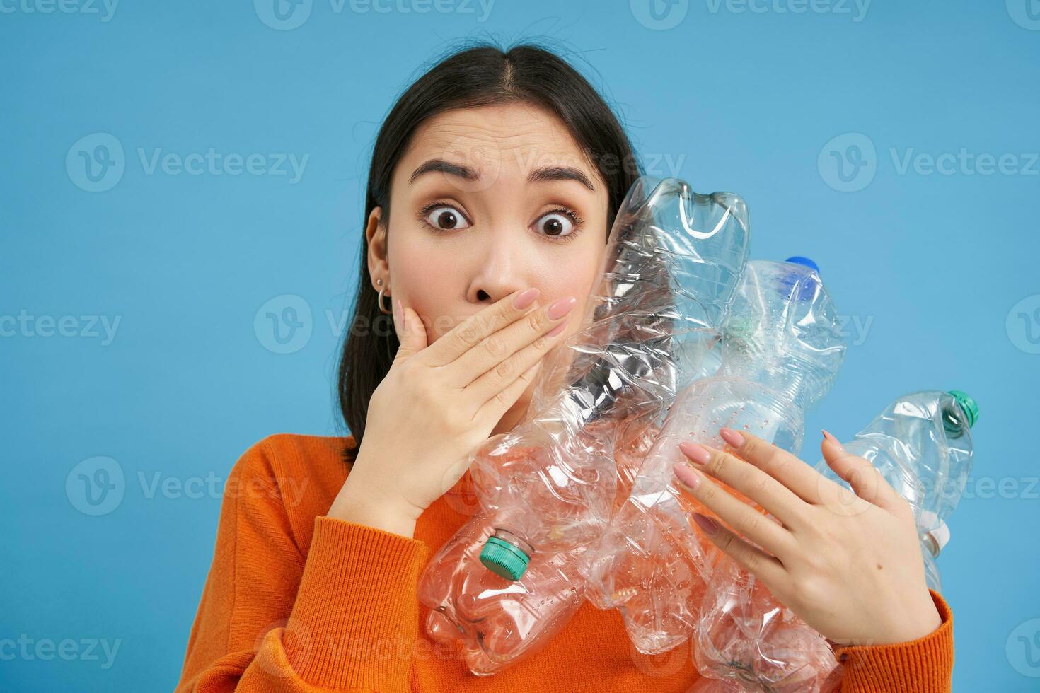 Surprised korean woman, holding plastic bottles, looking shocked at camera, concept of environment and recycling, blue background photo