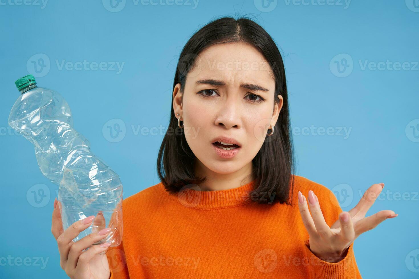 Portrait of confused woman, holding and showing plastic bottle, clueless why people are not recycling, blue background photo