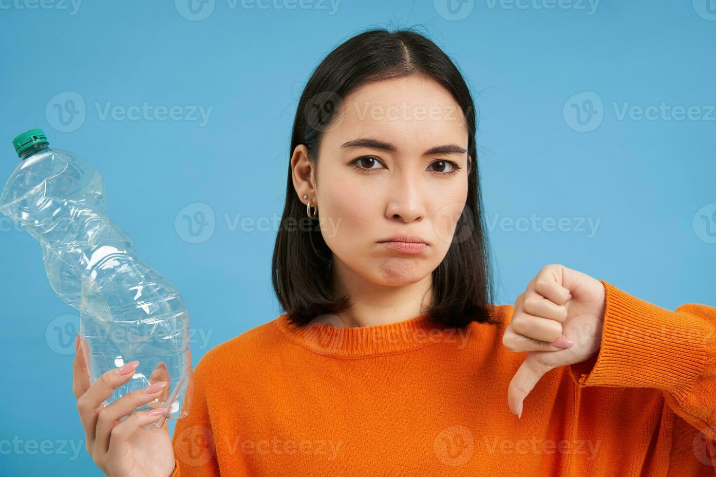 Portrait of asian woman shows thumbs down and plastic bottke, disapprove lack of recycling, blue background photo