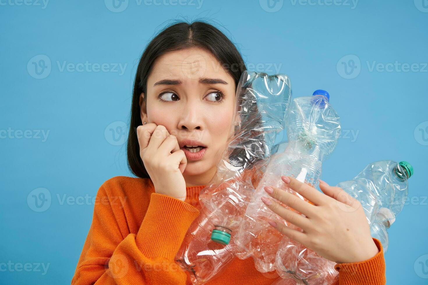 Portrait of asian girl with scared face, holding piles of plastic bottles for recycling and stares to the right with shocked emotion, blue backgrond photo