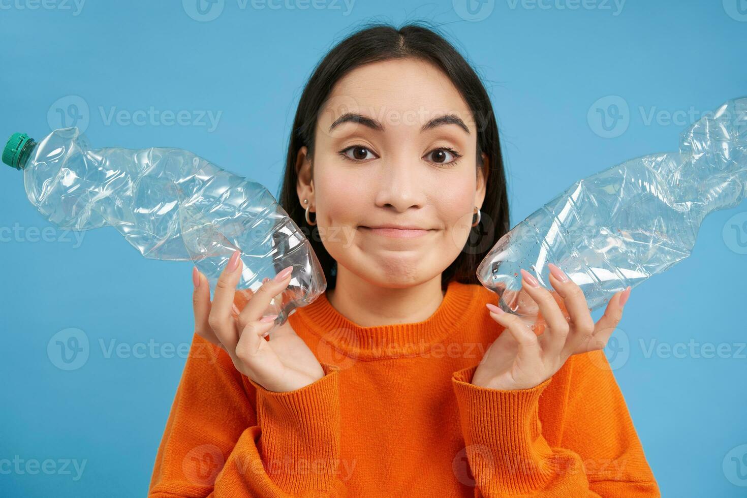 Cute korean woman shows two plastic bottles and smiles, recycles, sorts trash, collecting recycable items, blue background photo