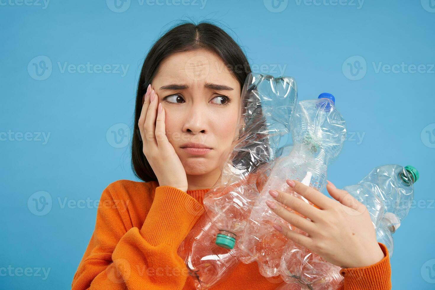 Sad asian woman holding plastic bottles, looking troubled and upset, sorting waste for recycling, standing over blue background photo