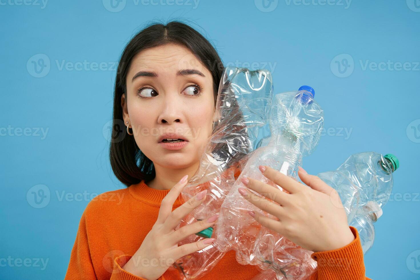 Portrait of asian girl with scared face, holding piles of plastic bottles for recycling and stares to the right with shocked emotion, blue backgrond photo