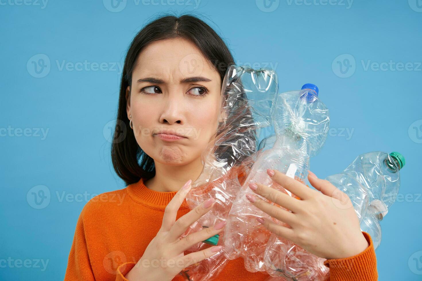 Annoyed korean woman looking with blaming face, holding plastic bottles, standing over blue studio background photo