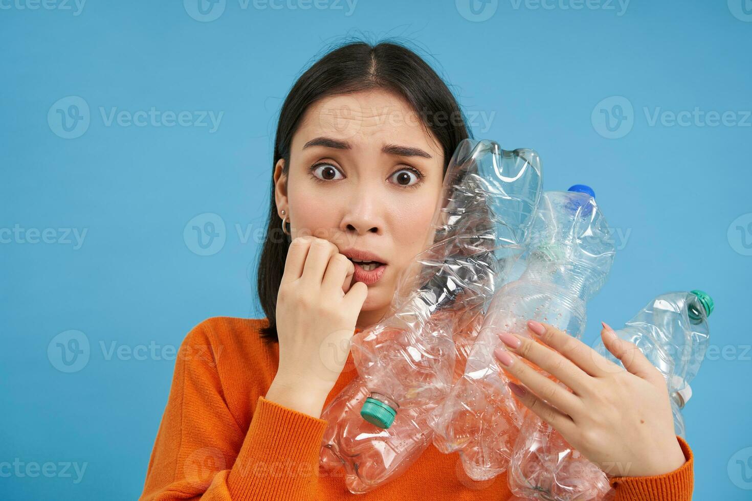 Surprised korean woman, holding plastic bottles, looking shocked at camera, concept of environment and recycling, blue background photo