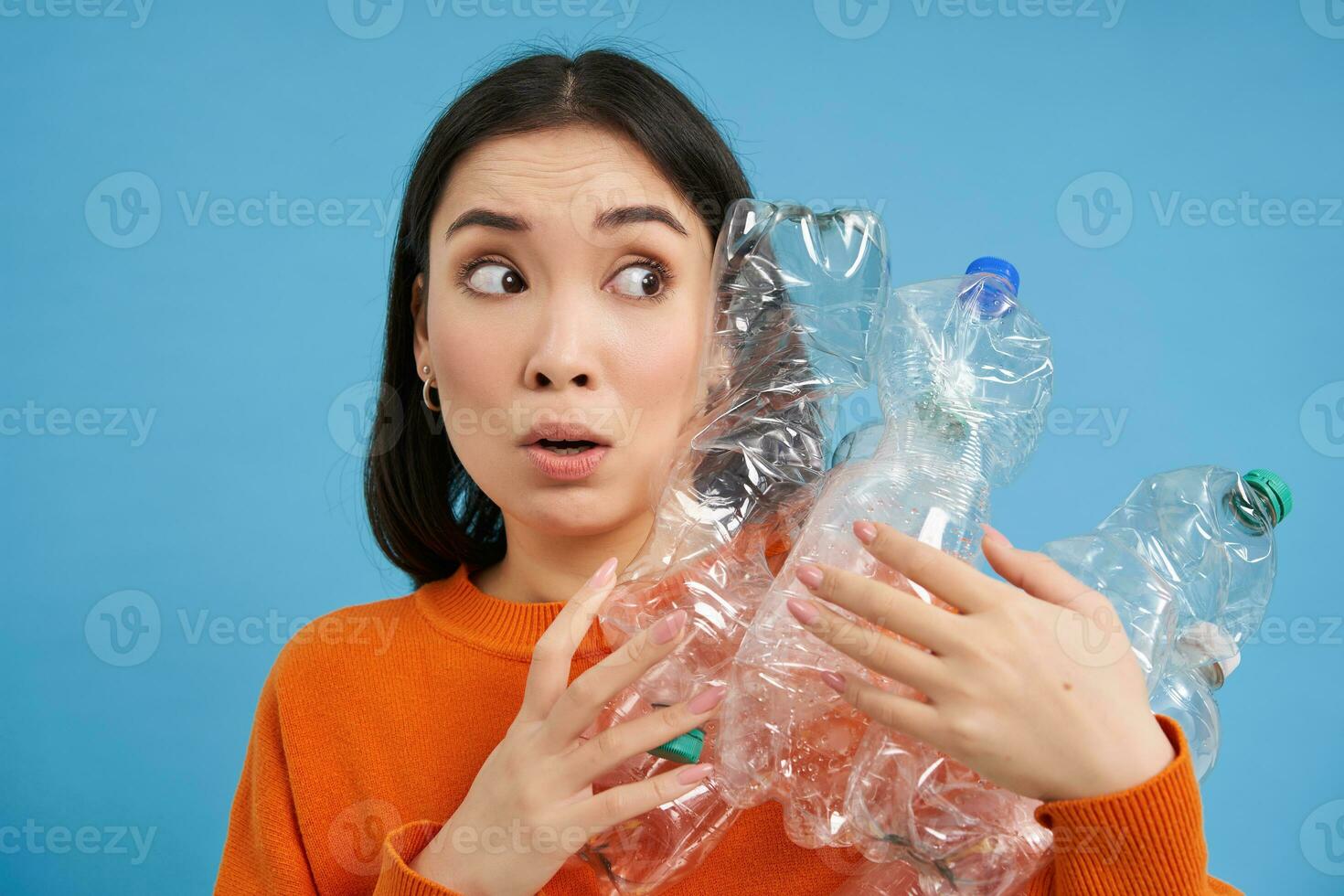Portrait of asian girl with scared face, holding piles of plastic bottles for recycling and stares to the right with shocked emotion, blue backgrond photo