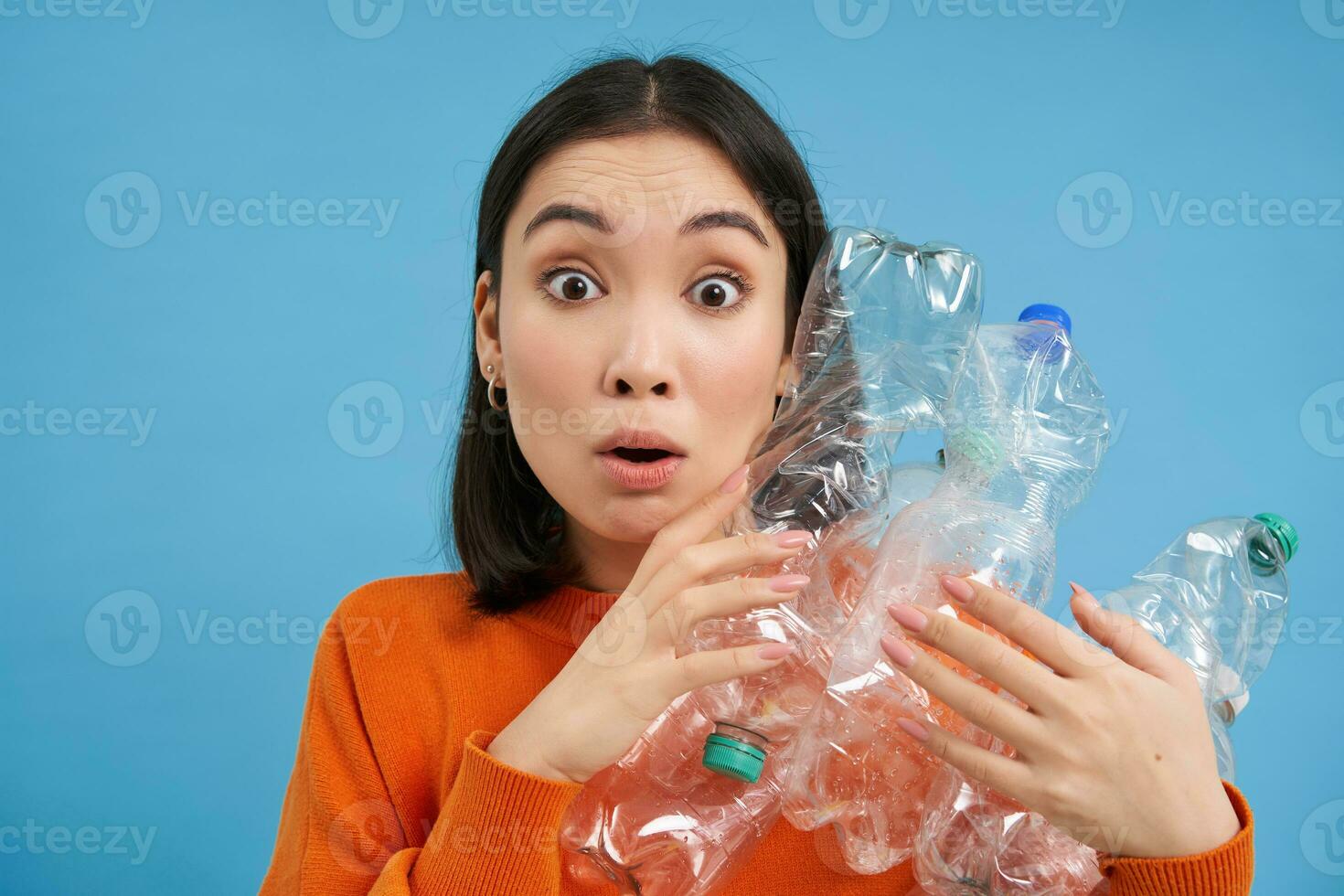 Surprised korean woman, holding plastic bottles, looking shocked at camera, concept of environment and recycling, blue background photo